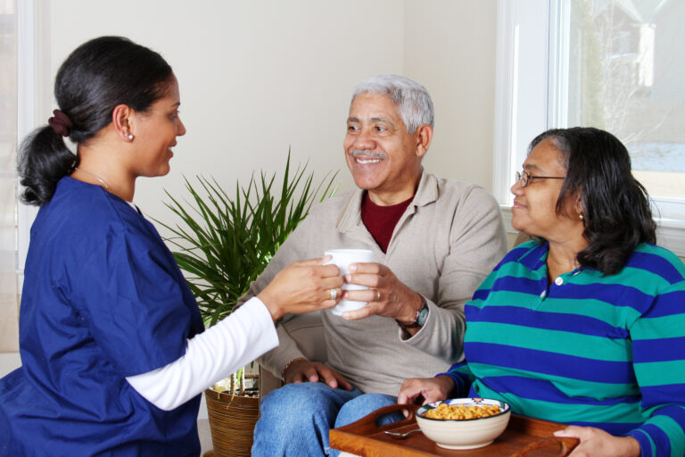 A senior companion caregiver serves lunch to an elderly couple sitting on the couch.